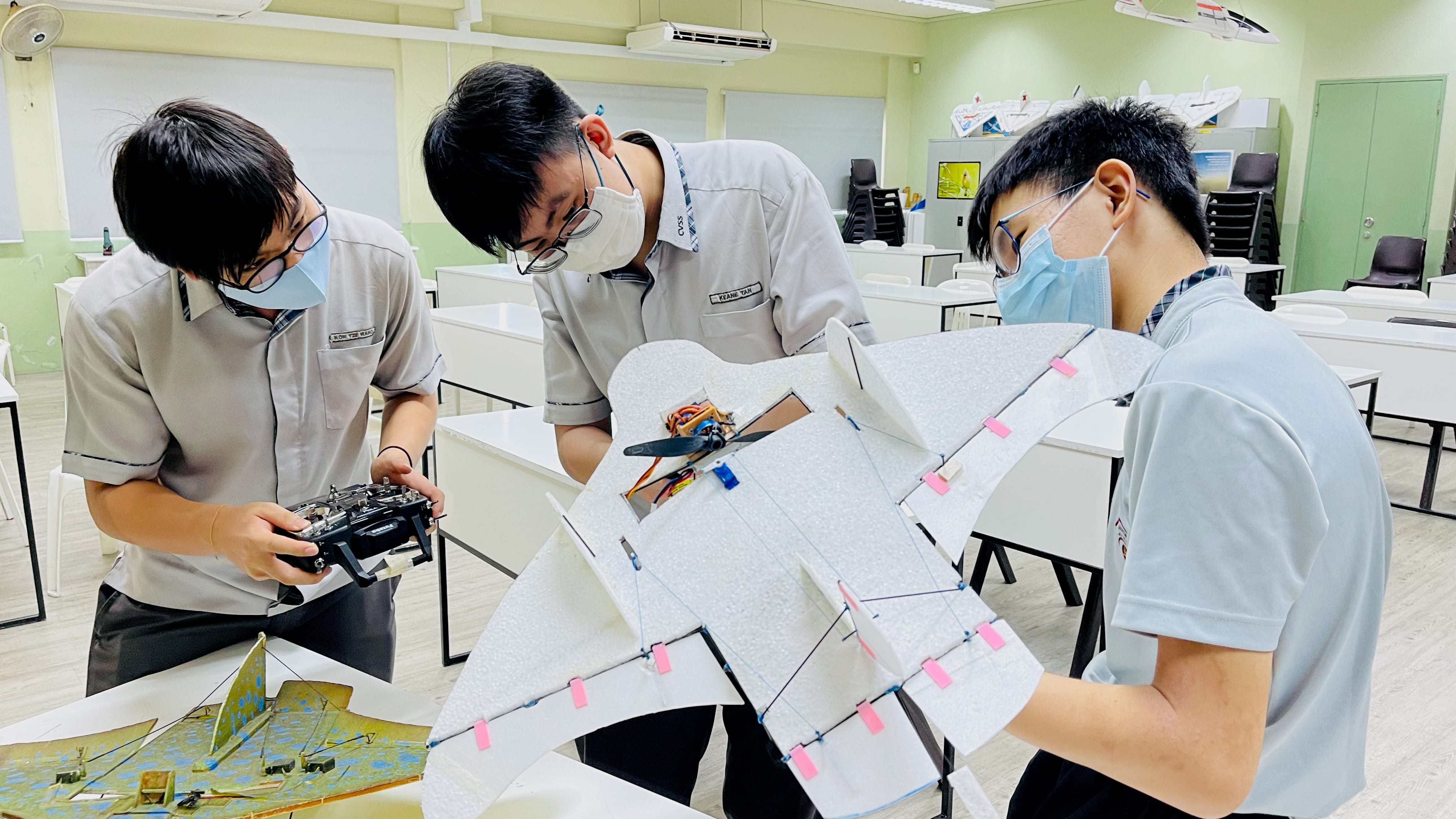 Three masked students in a classroom inspect a foam model plane while one holds a remote controller.